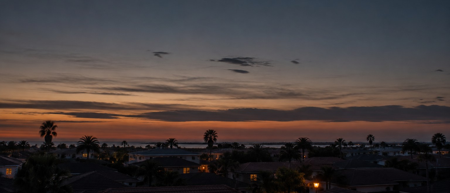 Dusk over a Gulf coast neighborhood, with tile rooftops and palms silhouetted against an orange and blue sky.